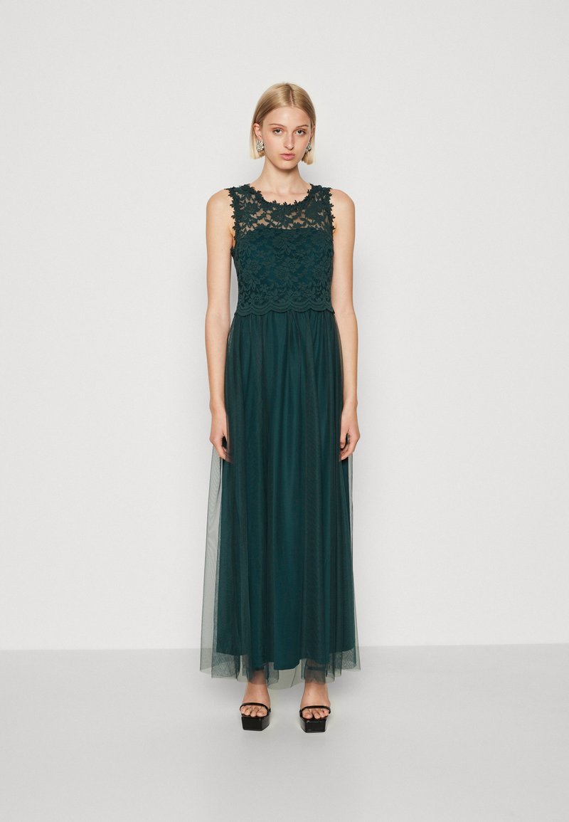 Rochie lungă de un verde închis, cu un corset din dantelă, design fără mâneci și fustă din tul fluidă. Asortată cu sandale cu platformă negre.