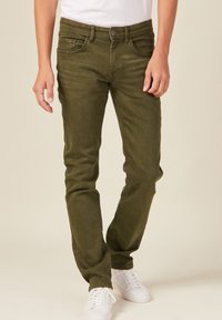 Jeans verde oliva de mezclilla, con un diseño clásico de cinco bolsillos, corte ajustado y cremallera con cierre de botón. Combinados con zapatillas blancas.