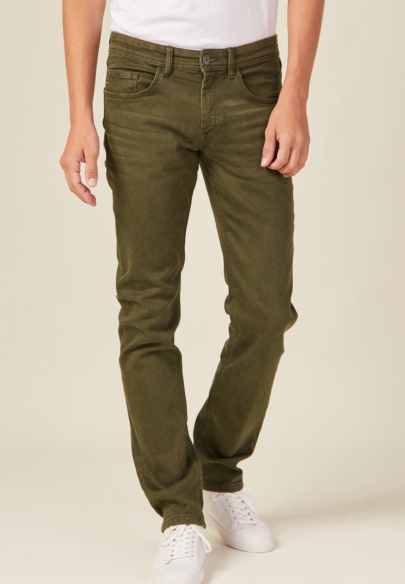 Jeans verde oliva de mezclilla, con un diseño clásico de cinco bolsillos, corte ajustado y cremallera con cierre de botón. Combinados con zapatillas blancas.