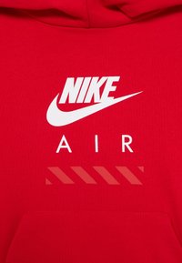 Crvena dukserica s kapuljačom koja ima bijeli Nike logo i tekst "AIR", uz prugaste naglaske dolje. Izrađena od mekog materijala, s prednjim džepom i vezicom za kapuljaču.