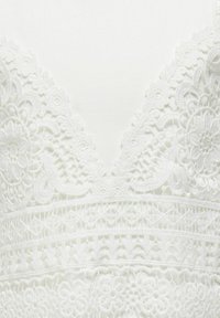 Hvid blondekjole med en dyb V-udskæring, indviklede blomster mønstre og strukturerede horisontale bånd. Blødt stof med lagdetaljer.