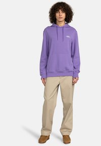 Felpa viola con tasca a canguro e cappuccio con coulisse, abbinata a pantaloni beige e scarpe marroni. Tessuto liscio e vestibilità casual.