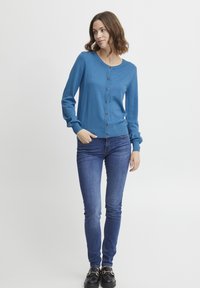 Cardigan albastru tricotat, cu guler rotund, mâneci lungi și nasturi din lemn pe față. Asortat cu blugi skinny și loafers negri.