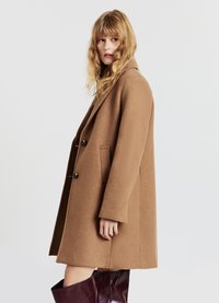 Cappotto di lana marrone con vestibilità rilassata, ampie rever, due bottoni neri e tasche laterali. Il tessuto testurizzato aumenta l'interesse visivo.