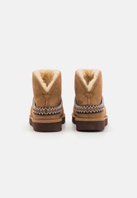 Brune suede ankelstøvler med blød pelsfor, der har et dekorativt vævet bånd omkring mellemsålen og en flad gummisål.