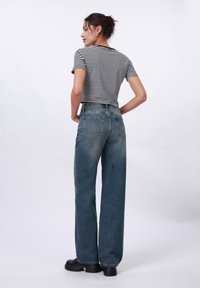 Ženska stoji obrnjena stran, oblečena v črno-belo prugasto majico s kratkimi rokavi, visoko pasne široke modre kavbojke in črne platforme.