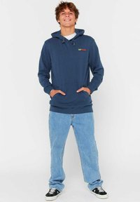 Granatowa bluza z kapturem, ze sznurkiem i kieszenią kangurką, zestawiona z jasnoniebieskimi spodniami w prostym kroju i czarnymi sneakersami. Logo "RIP CURL" na klatce piersiowej.