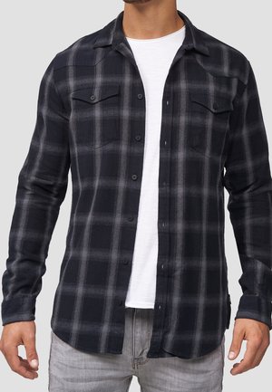 Schwarz-grau karierter Flanellhemd mit Knopfleiste, zwei Brusttaschen und langen Ärmeln. Über einem weißen T-Shirt getragen und mit hellgrauen Jeans kombiniert.