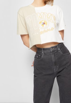 Osoba nosząca krótką, dwukolorową beżowo-białą koszulkę Tommy Jeans z żółto-białym logo oraz ciemnoszare jeansy z wysokim stanem.