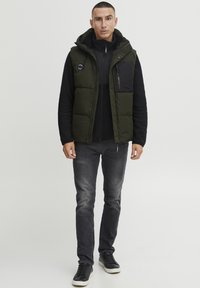 Grüne Steppweste mit Kapuze, schwarzen Akzenten und einer Reißverschlusstasche, getragen über einem schwarzen Reißverschlussoberteil, kombiniert mit grauen Jeans und schwarzen Sneakers.