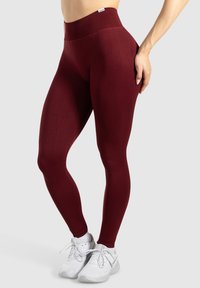 Leggings bordeaux con vita alta, realizzati in tessuto elasticizzato, con una texture liscia e un design aderente. Indossati con scarpe da ginnastica bianche.