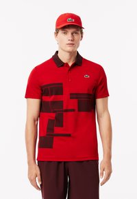 Röd polo med mörkbruna geometriska mönster, korta ärmar och krage. Matchad med en röd keps med en logotyp.