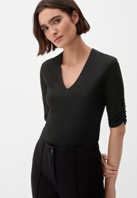 Zwarte aansluitende catsuit met een V-hals en driekwart mouwen, voorzien van geplooide details op de mouwen en een zachte textuur.