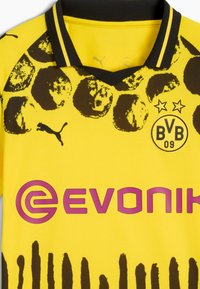 Camiseta de fútbol amarilla con acentos negros, cuello en V y un gráfico de balones de fútbol; logotipos en rosa y negro a la altura del pecho.
