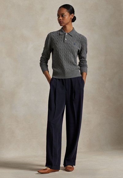 Nainen seisoo kädet taskuissa, päällään harmaa palmikkoneulepikeepaita, leveät tummansiniset housut ja ruskeat tasapohjaiset kengät, neutraali tausta.