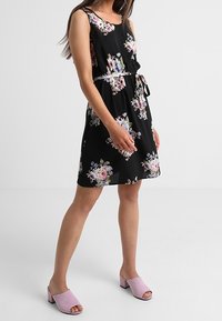 Černé květinové šaty se bezrukávovým designem, páskem v oblasti pasu a splývavým střihem. V kombinaci s růžovými otevřenými pantoflemi.