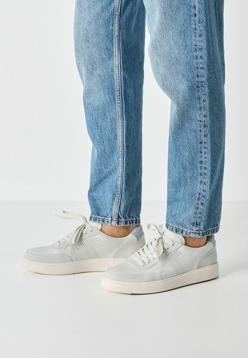 Ανοιχτό γκρι δερμάτινα sneakers με διάτρητες λεπτομέρειες, επίπεδα κορδόνια και υφασμένο λευκό λαστιχένιο πέλμα, συνδυασμένα με χαλαρά μπλε τζιν jeans.