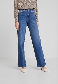 Jeans de pierna ancha azul claro con un lavado medio. Presentan un diseño clásico de cinco bolsillos y detalles de costura. Llevados con una blusa de estampado de leopardo.