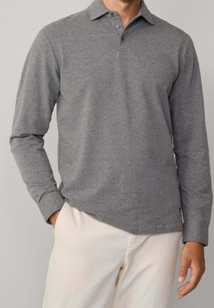 Mand iført en grå poloshirt med lange ærmer og to-knaps lukning samt lyse beige bukser, stående med den ene hånd i lommen.