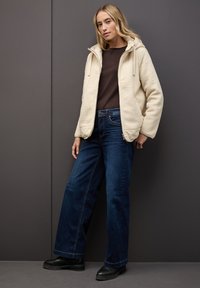 Chaqueta de forro polar marfil con capucha, llevada sobre una blusa marrón, combinada con unos vaqueros de pierna ancha azul oscuro y botas de plataforma negras. Fondo neutro.