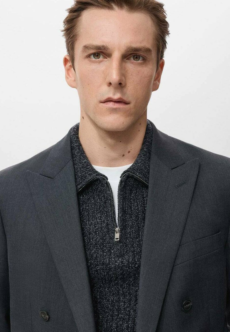 Blazer grigio doppiopetto indossato sopra un maglione scuro a maglia. Il blazer presenta rever a lancia e decorazioni minime. Design semplice e sartoriale.