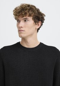 Melna adīta džemperis ar apaļu kaklu, ar faktūru rakstu un ribotām piedurknēm. Modelim ir čirkaini mati un neitrāla izteiksme.