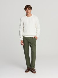 Biały sweter z dzianiny warkoczowej, oliwkowe spodnie cargo z bocznymi kieszeniami, kremowe podeszwy, model stojący na jednolitym tle.