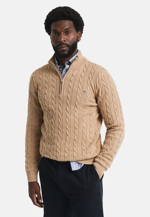 Beige kabelgebreide trui met een hoge kraag en kwart rits. Kenmerkt zich door geribbelde manchetten en zoom, gedragen over een lichtblauw shirt.