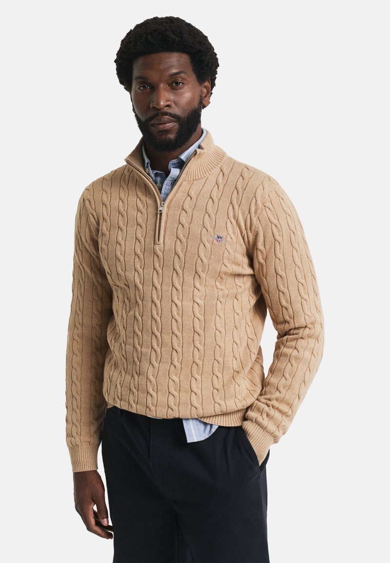 Beige Strickpullover mit hohem Kragen und Viertelreißverschluss. Mit gerippten Bündchen und Saum, getragen über einem hellblauen Hemd.