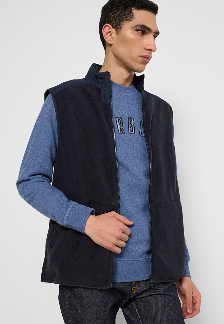 Gilet en polaire marine avec un col haut et des poches avant, superposé sur un sweat-shirt bleu. Doté d'une fermeture éclair et d'une texture légère.