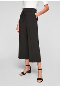 Zwarte brede pijpen met een hoge taille, voorzien van zijzakken en een trekkoorddetail, gemaakt van een soepele stof. Gecombineerd met zwarte hakken met enkelbandje.