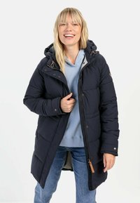 Marineblå pufferjakke med hætte, med lynlås- og knaplukning, quiltet design og sidelommer. Bæres over en lys blå hoodie.