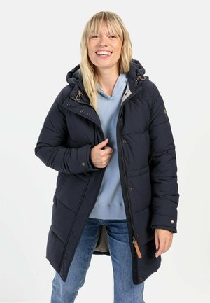 Donkerblauwe poffermantel met een capuchon, featuring een ritssluiting en knoopsluiting, geborduurd design en zijzakken. Gede gedragen over een lichtblauwe hoodie.