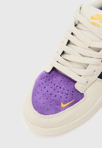 Des sneakers Nike avec une tige en daim beige, un bout en daim violet, des accents orange et une semelle en caoutchouc blanc. Les lacets sont de couleur beige clair avec une languette en mesh.