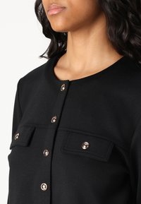 Schwarzes Stofftop mit rundem Halsausschnitt, goldfarbenen Druckknöpfen und zwei vorderen Taschen mit Klappen. Glatte Textur mit taillierter Passform.