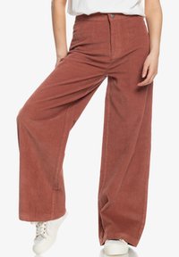 Pantalons en velours côtelé à jambes larges de couleur rouille avec des côtes verticales, taille haute et fermeture à bouton, associés à des baskets blanches.