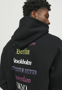 Sudadera negra con gran texto impreso y colorido en la parte posterior que presenta nombres de ciudades. El material parece suave, con una capucha amplia y un corte relajado.