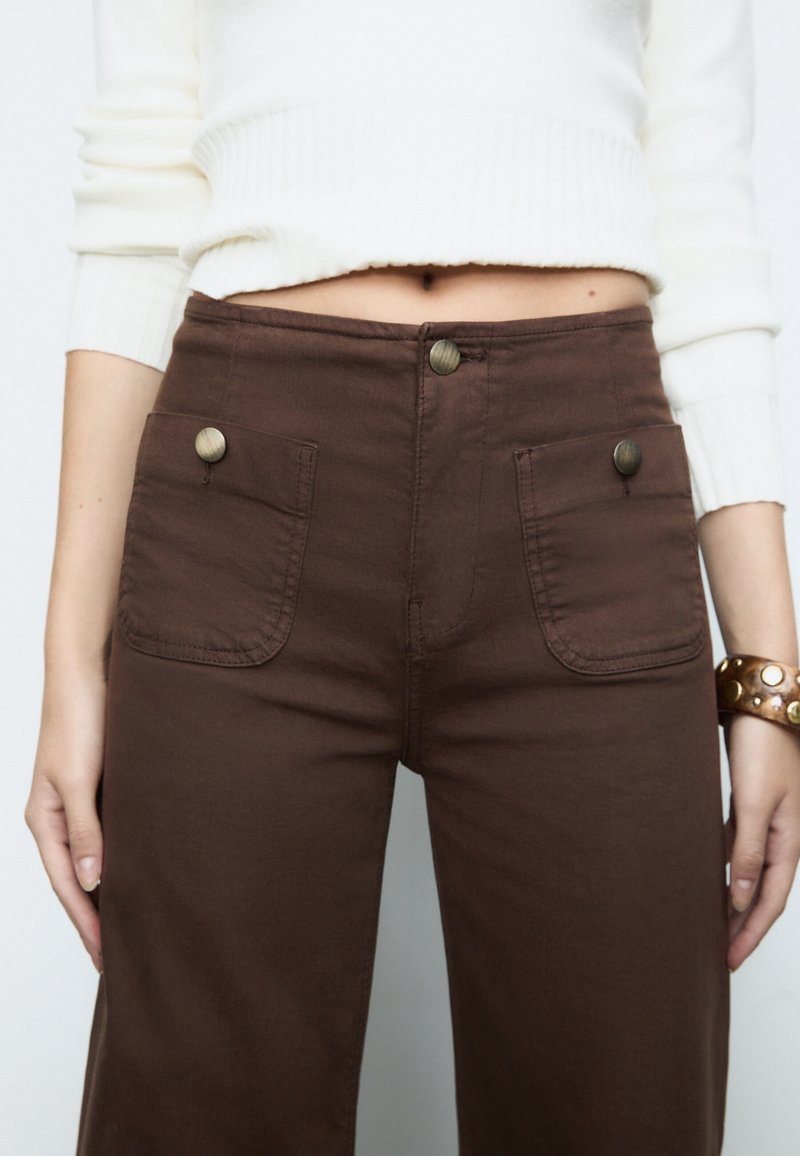 Pantalon marron taille haute avec poches plaquées à l'avant et boutons en laiton, porté avec un pull court blanc côtelé et un bracelet en cuir clouté.
