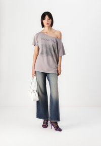 Grå oversized t-skjorte med "New York" grafikk, parret med falmede, vide denimjeans og lilla stroppehæler. Hvit veske som accent.