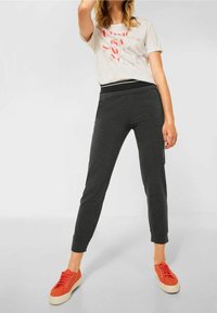 Grå mjukisbyxor med svart elastisk midja, kombinerat med en ljus grafisk t-shirt och röda sneakers, stående i en uppställd pose.