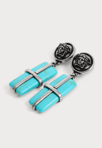 Boucles d’oreilles pendantes en argent avec des médaillons circulaires présentant un visage et des pierres turquoise rectangulaires maintenues par des bandes en argent.