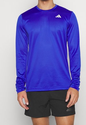 Mies, joka pukeutunut kirkkaan siniseen pitkähihaiseen urheilupaitaan ja mustiin shortseihin, seisoo yksinkertaisen valkoisen taustan edessä.