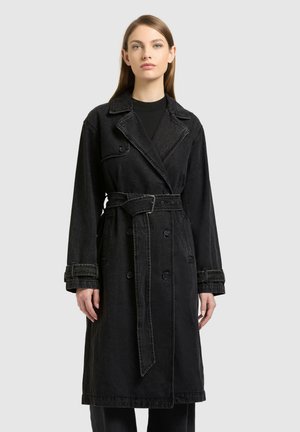 Žena stojí oblečená v dlhom čiernom rifľovom trenčkotom s opaskom, širokými klopami a zapínanými manžetami, na jednotnom svetlom pozadí.