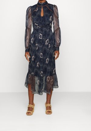 Granatowa sukienka midi w kwiatowe wzory, przezroczyste pękate długie rękawy, dekolt w kształcie klucza, falbaniasty dół, noszona z beżowymi sandałami na obcasie z paskami.