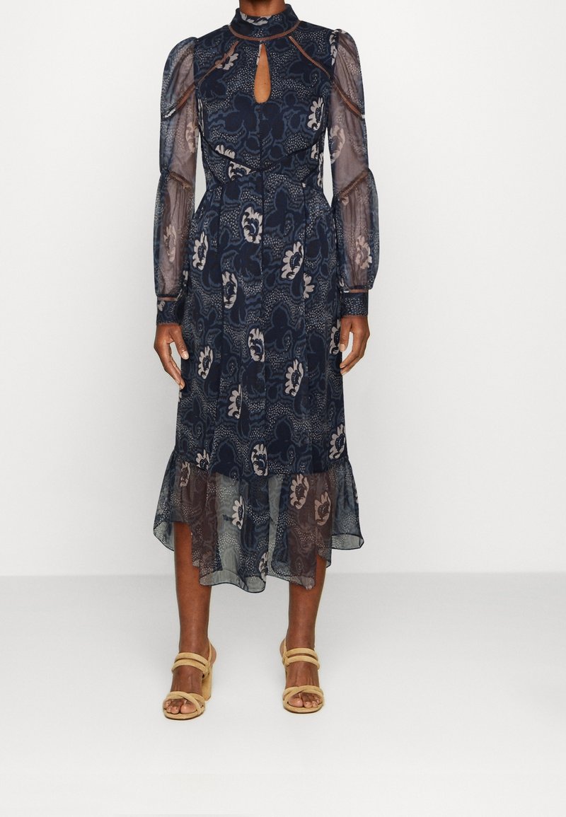 Robe midi bleu marine à motifs floraux, manches longues transparentes bouffantes, encolure en trou de serrure, ourlet froufroutant, portée avec des sandales beiges à talons et brides.