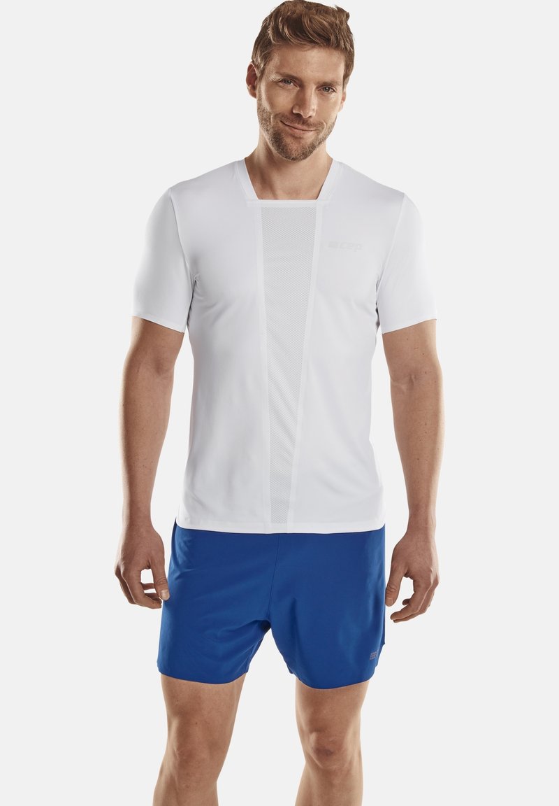 Weißes sportliches T-Shirt mit kurzen Ärmeln und Netz-Einsätzen, kombiniert mit blauen Shorts. Das Shirt hat eine angepasste Passform und eine leichte Textur.