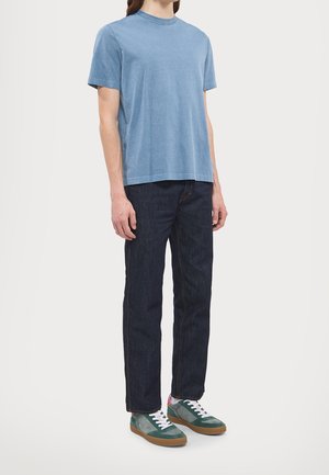 Homme portant un t-shirt bleu uni à manches courtes, un jean bleu foncé et des baskets vertes et blanches, debout devant un fond blanc.