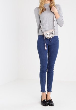 Donna con una maglietta a maniche lunghe grigia, jeans skinny blu, mocassini neri e una piccola borsa a tracolla con motivo pitonato.