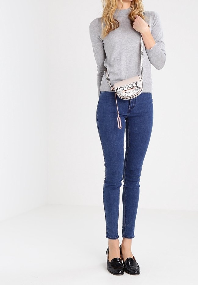 Mujer que lleva una camiseta de manga larga gris, jeans ajustados azules, mocasines negros y lleva un pequeño bolso cruzado con un patrón de piel de serpiente.