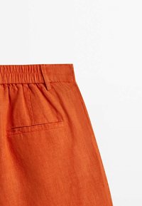 Gros plan sur un pantalon en lin orange avec une taille élastique, des passants de ceinture et une poche passepoilée à l'arrière du côté droit.